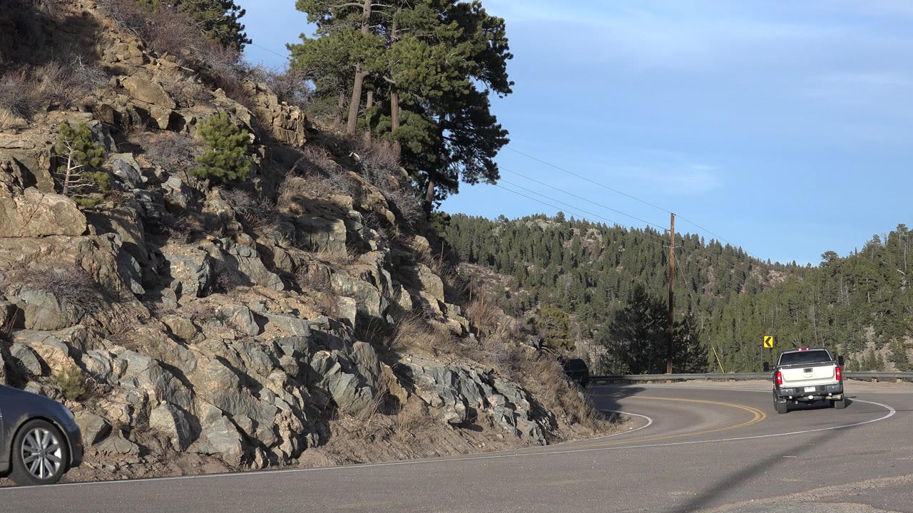 A white pickup moves away on the right side of the Nederland, Colorado road while the front of a dark car enters from the lower right, with rocks, pines, and blue sky visible, and no visible people.