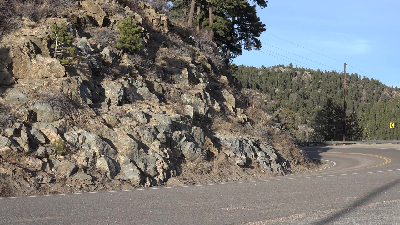 Opening frame of a Nederland, Colorado mountain road curving past a rocky roadside slope, with pine-covered hills, blue sky, and no visible people.