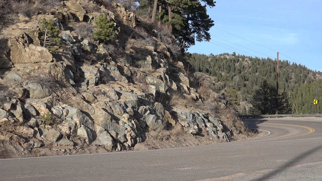 Sunny road bend below rock formations and pine trees.