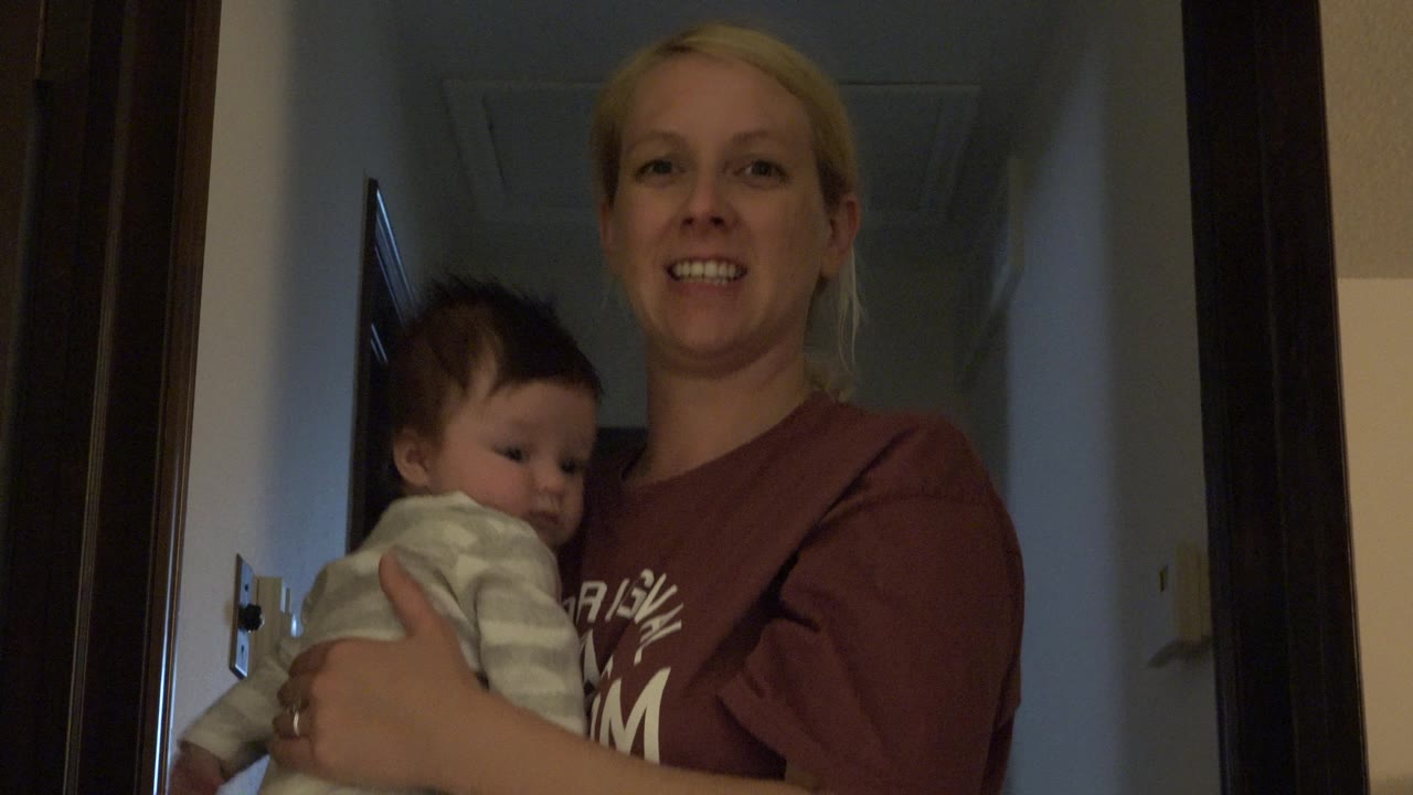 The woman smiles toward the camera while holding the newborn in the same narrow Westminster, Colorado hallway, with warm low indoor light on their faces.