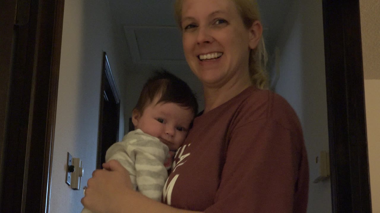 Blonde woman smiling while holding a baby indoors near a doorway.