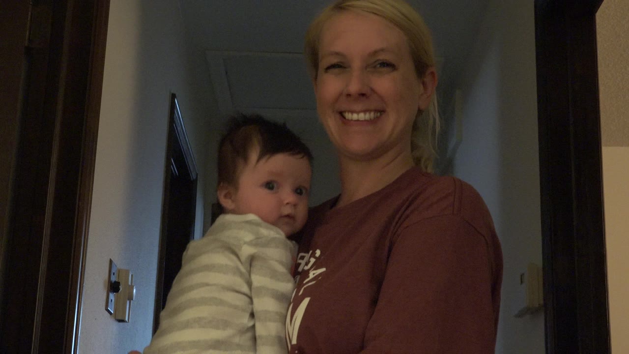 Blonde woman smiling while holding a dark-haired baby indoors.
