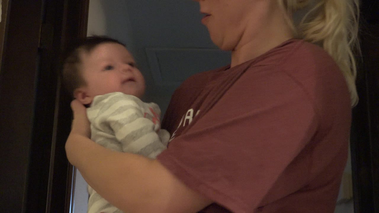 A blonde woman in a maroon shirt holds a baby in a gray striped outfit near a dark interior doorway in Westminster, Colorado.