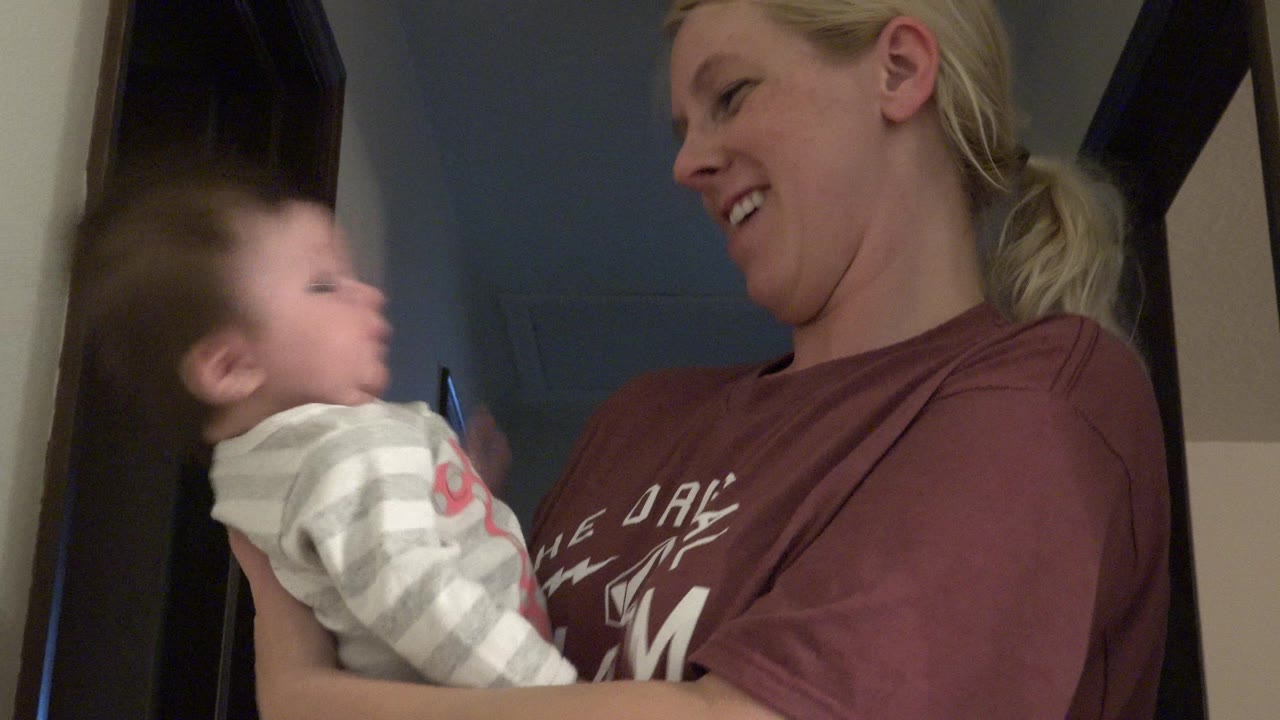 Blonde woman smiles while holding a baby indoors.