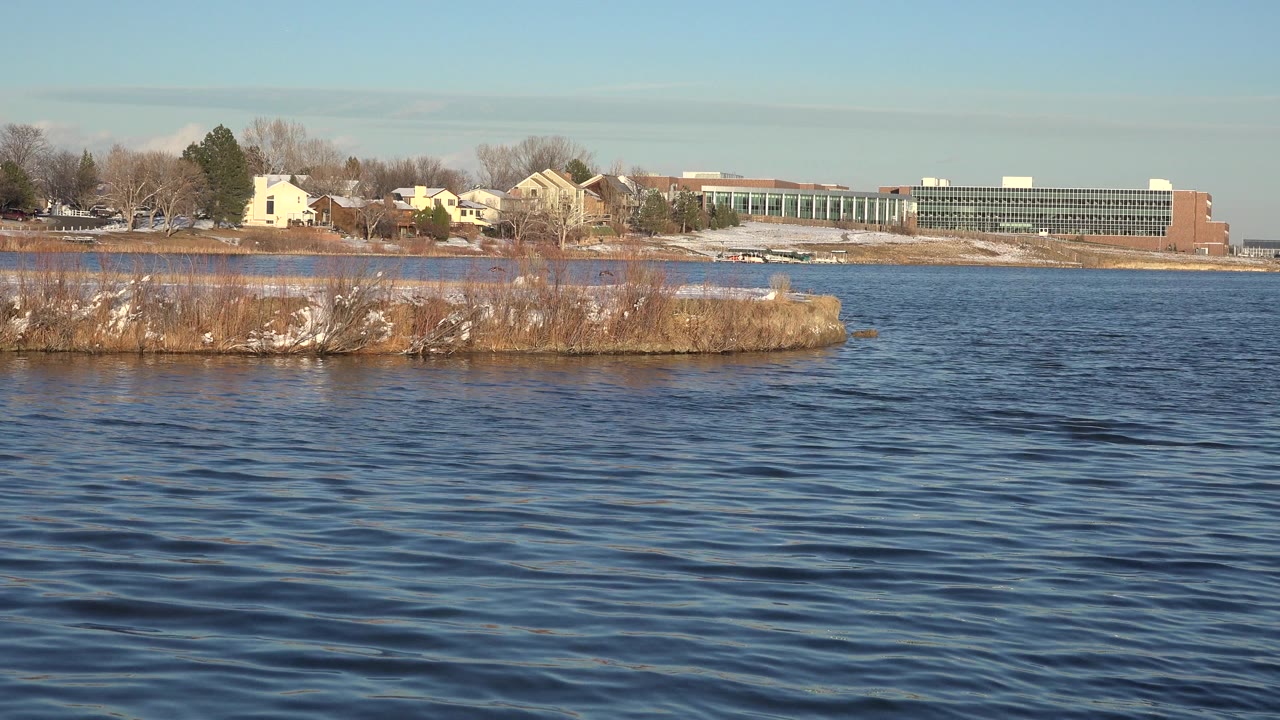 Second moment over blue water with the low Westminster shoreline running across frame, small snow patches on the bank, and no visible people.