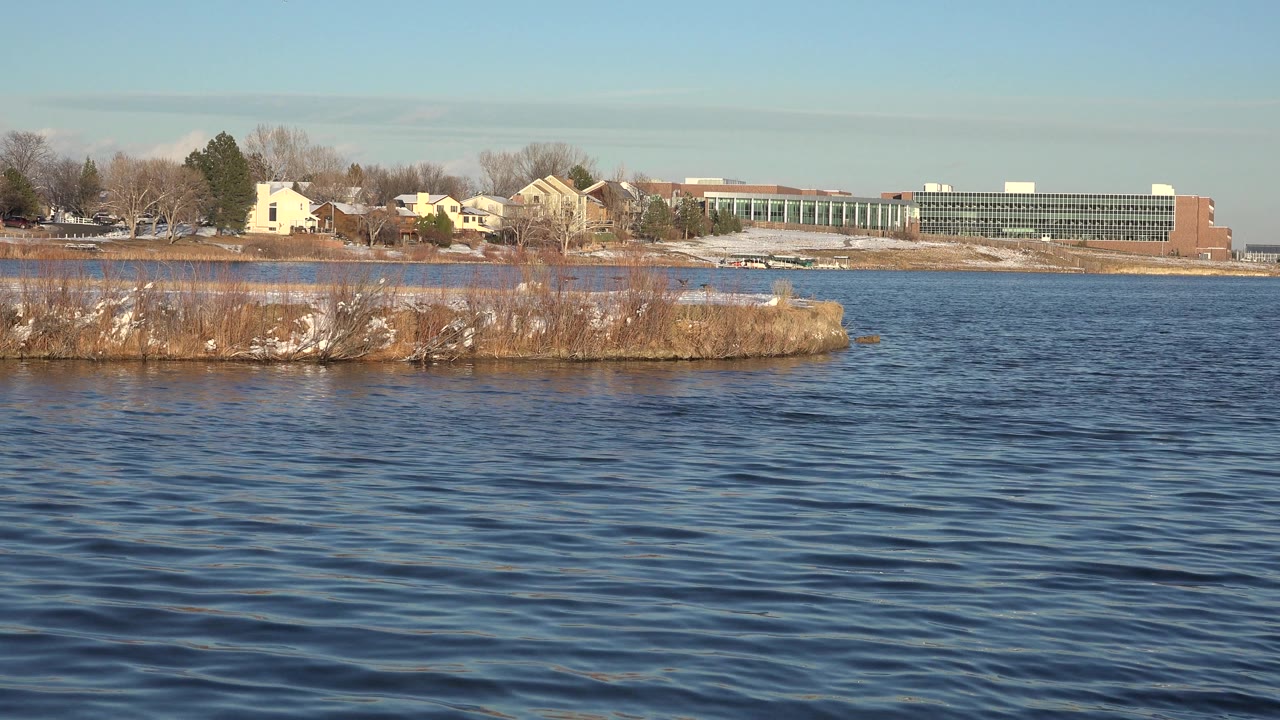 Wide water view with houses, bare trees, and a long building beyond the shore.
