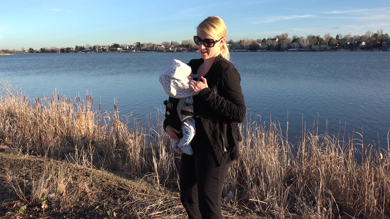 At the start of the Westminster, Colorado clip, a blonde woman in sunglasses holds a baby in white beside dry reeds and blue lake water.