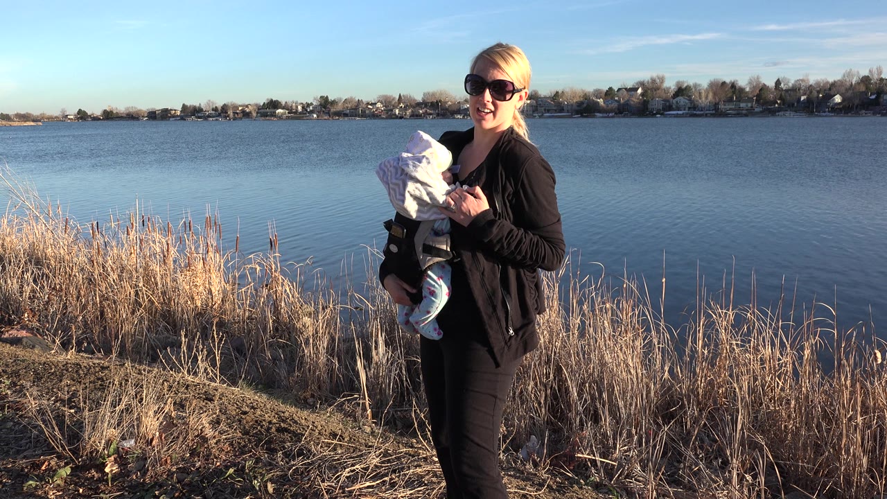 Woman faces camera while holding baby near a lake.