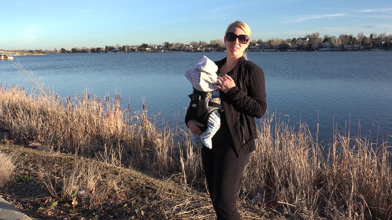 In Westminster, Colorado, a woman in dark clothing holds a bundled child near dry shoreline grasses with a blue lake and distant houses behind her.