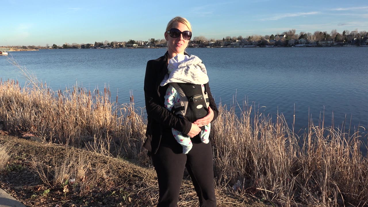 Woman wearing sunglasses holds a baby beside a lake with reeds in front.