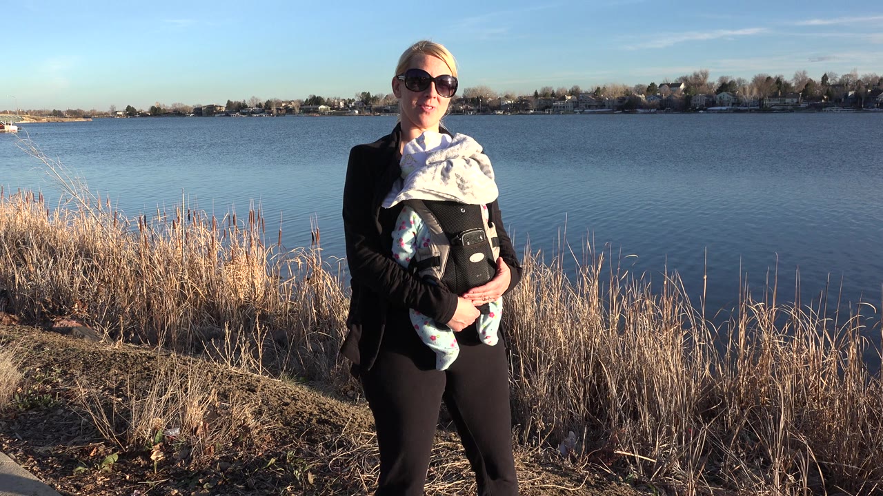 At 0:01, the woman stands more centered in the frame with the baby carrier visible against the lake and residential shoreline in Westminster, Colorado.