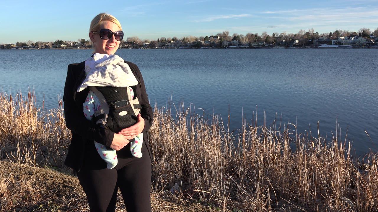 Blonde woman in sunglasses holding a baby beside a lake.