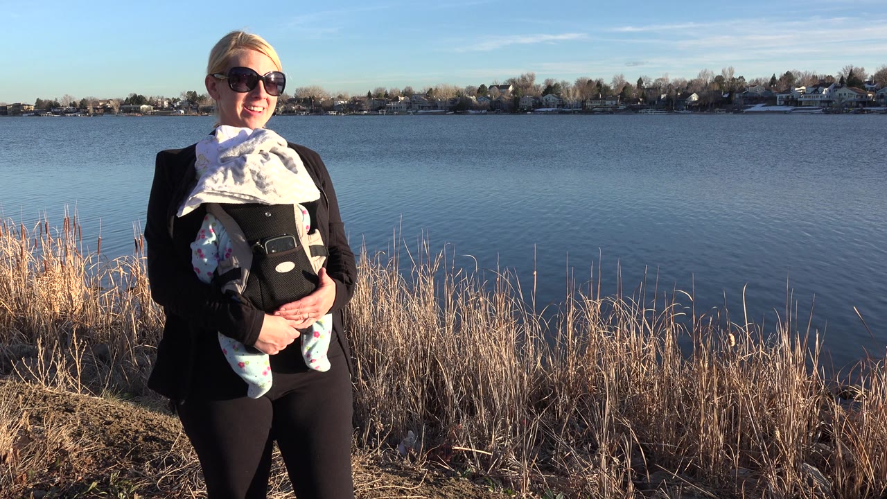 The woman and baby remain near the center of the Westminster lakeside frame, lit by strong sun with dry grasses in front and houses across the water.