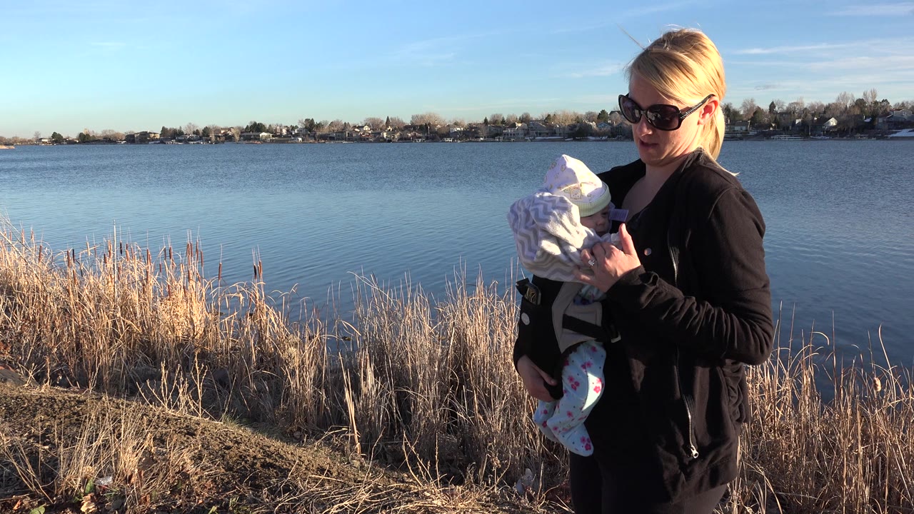 Woman holding a bundled baby beside a lake shore.