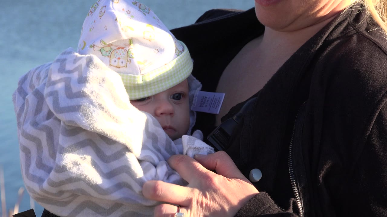 In Westminster, Colorado, the baby's eyes are visible between the blanket and cap while the woman supports the child against a blue water background.