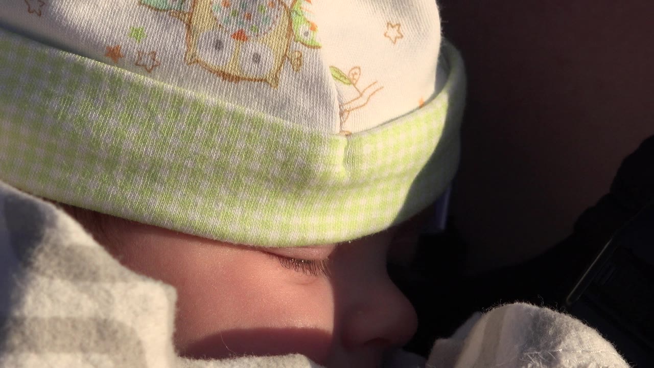 The baby remains in close-up in Westminster, Colorado, with the hat brim and gray blanket obscuring much of the face while sunlight falls across the cheek.