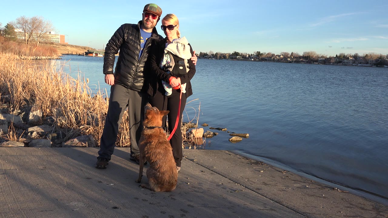 At the start of the Westminster, Colorado clip, a man and woman pose beside a lake while the woman holds a baby and a brown dog sits in the foreground on a red leash.
