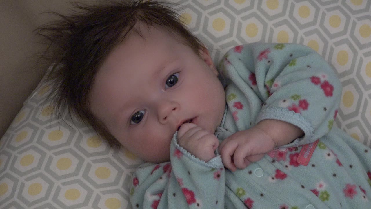 At 0:00, a baby in a pale blue sleeper lies on a gray and yellow patterned cushion in Westminster, Colorado, with one hand near the mouth and eyes looking toward camera.