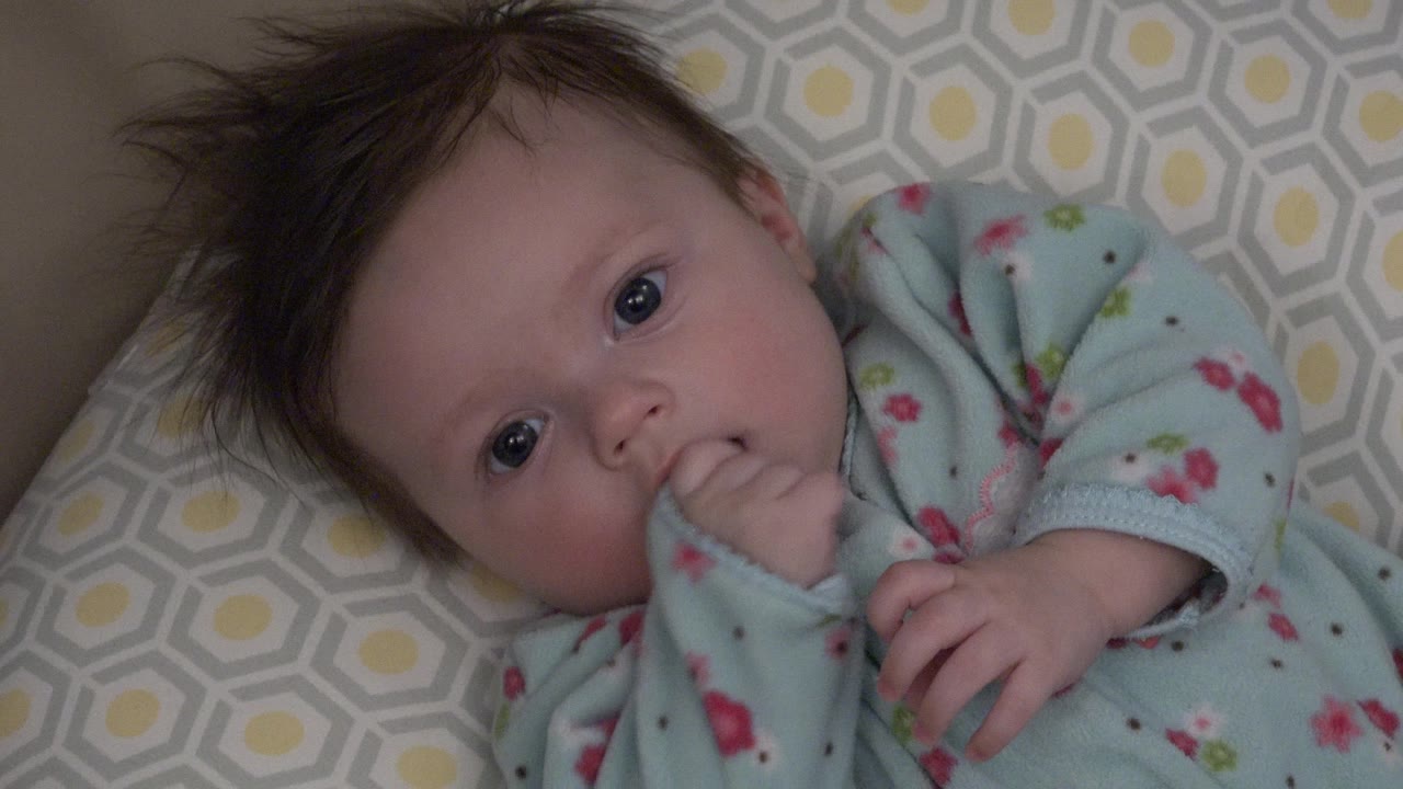 In Westminster, Colorado, a baby in a light blue floral sleeper lies on gray and yellow patterned bedding with one hand touching the mouth at the start of the clip.