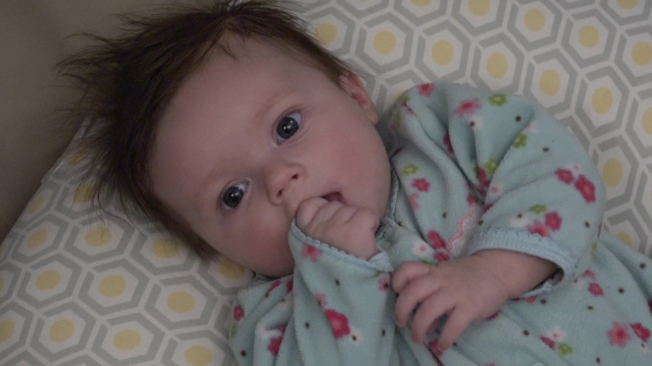 In Westminster, Colorado, the baby keeps fingers in the mouth while lying diagonally across the gray geometric cushion with yellow accents visible behind the head.