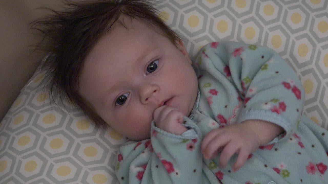 At 0:00, a baby in Westminster, Colorado lies close to camera on a gray and yellow patterned cushion with tousled hair and one hand near the mouth.
