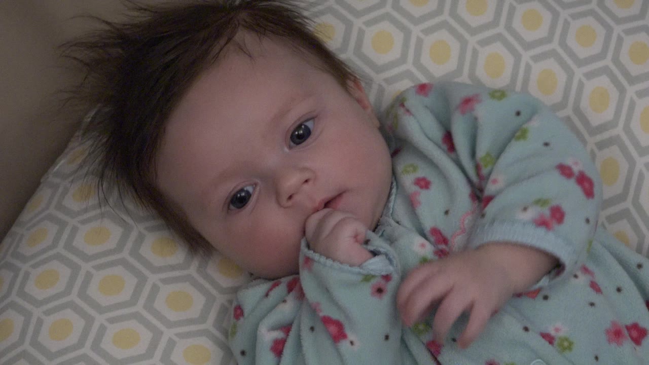 At 0:01, the baby looks toward the camera with wide eyes while chewing on a hand, with the pale blue floral sleeper visible against the patterned cushion.