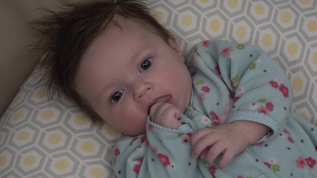 Baby with raised hand and fingers near the mouth on a patterned cushion.