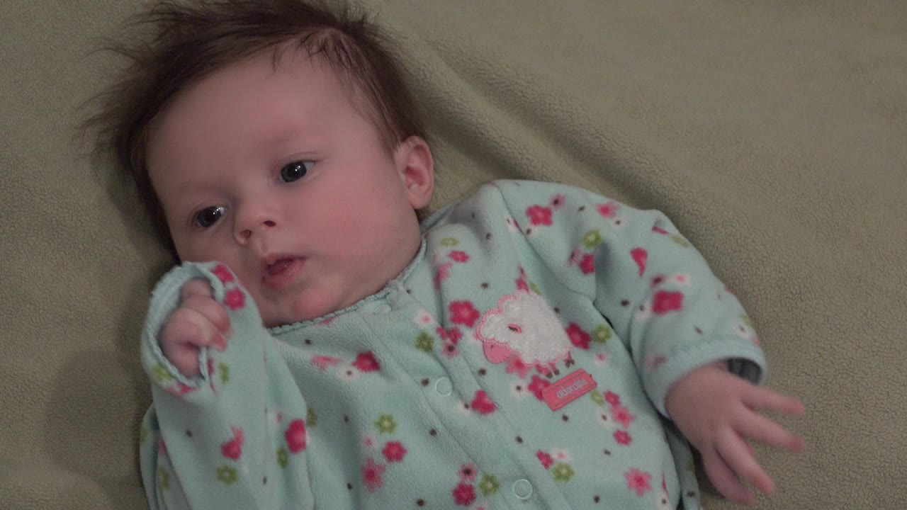 At 0:00, a baby in a mint flowery sleeper lies on a green blanket in Westminster, Colorado, with one hand near the face and the other arm extended.