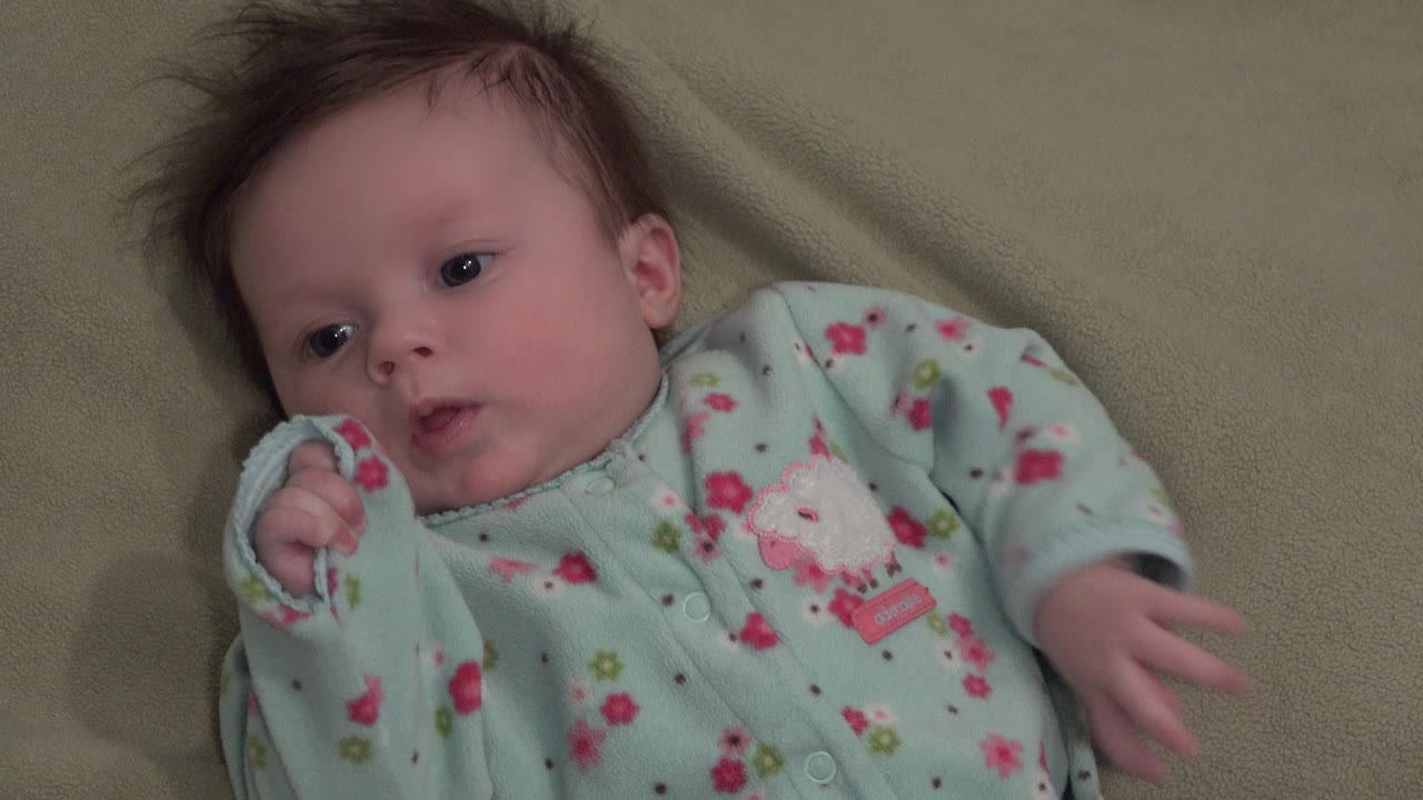At 0:01, the baby remains on the green blanket in Westminster, Colorado, looking upward while a covered hand rests close to the mouth.
