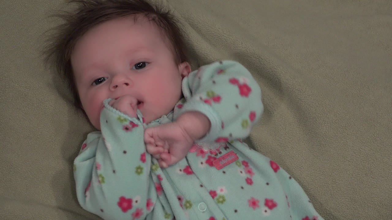 Infant girl lying on a beige blanket with fingers near her mouth.
