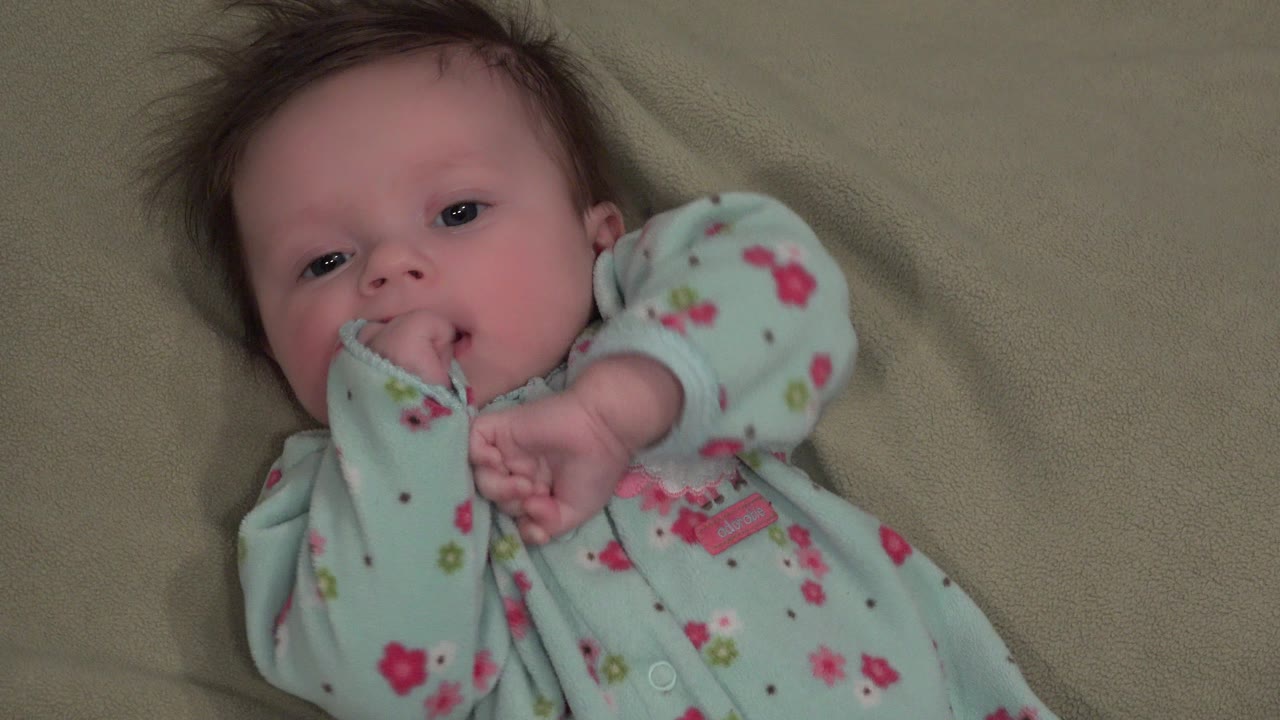 At 0:01, the infant girl remains on the beige blanket with her sleeve drawn to her lips and her other hand raised in front of her chest.