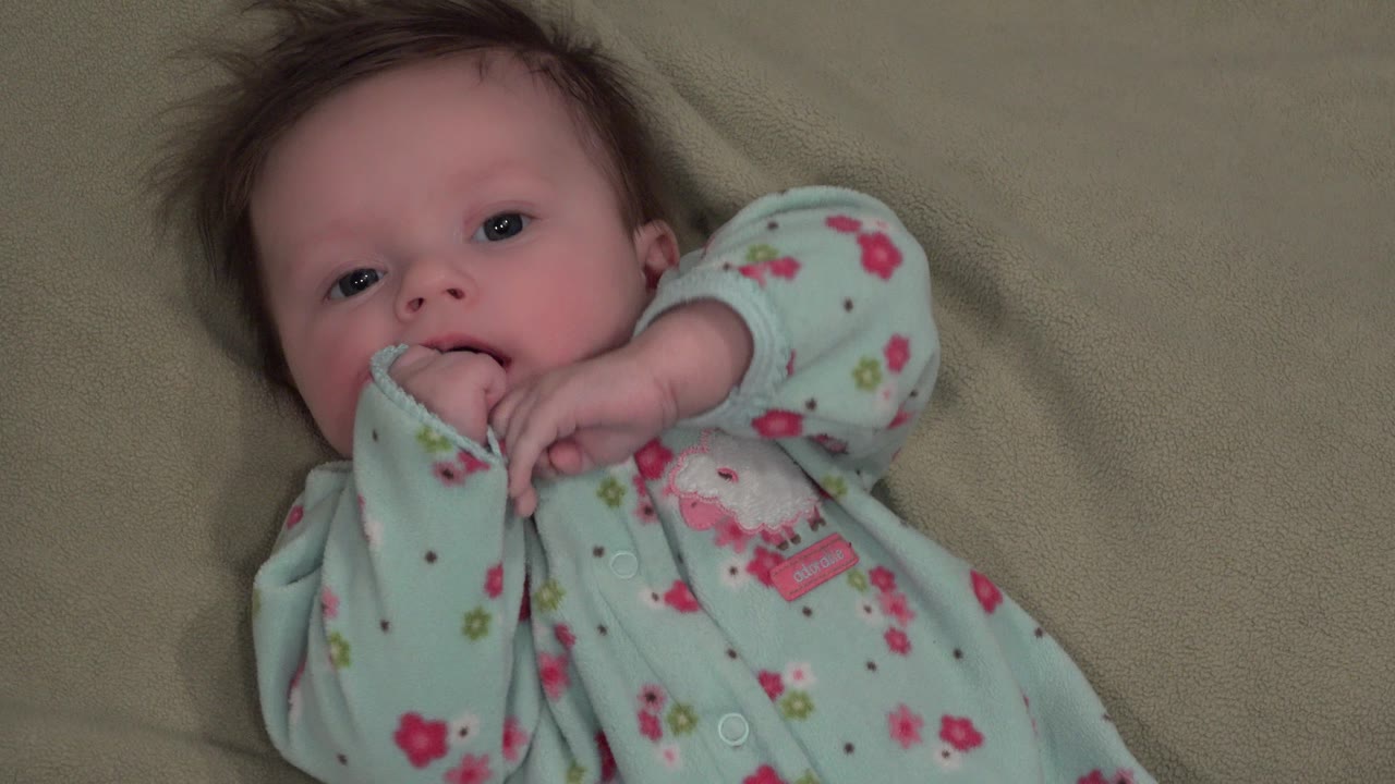 At 0:02, the baby in Westminster, Colorado has her fingers at her mouth while one bent arm crosses the foreground of the close indoor frame.