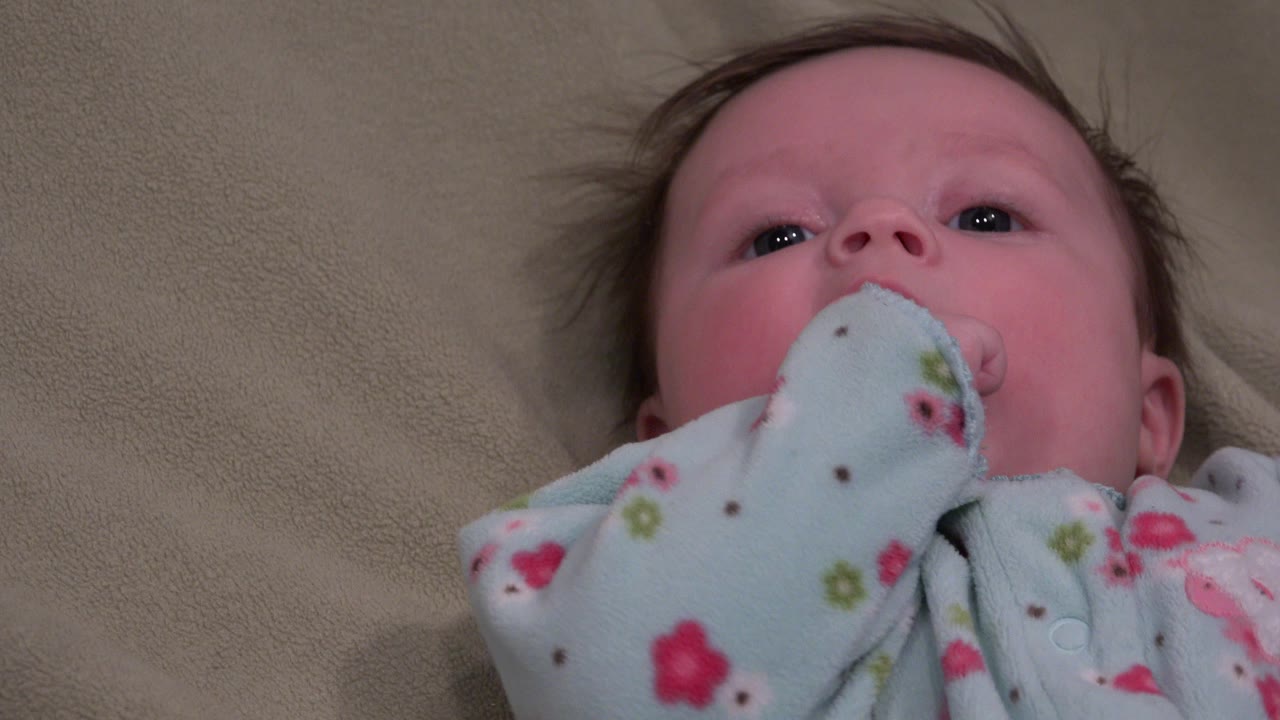 Baby in floral pajamas lying on beige bedding with sleeve at mouth.