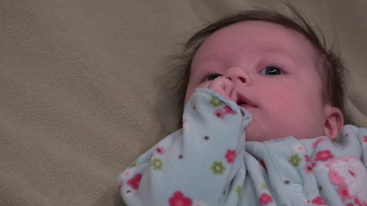 A baby in Westminster, Colorado lies on a tan surface with wide eyes, flyaway hair, and a pale blue floral sleeve raised to the mouth.