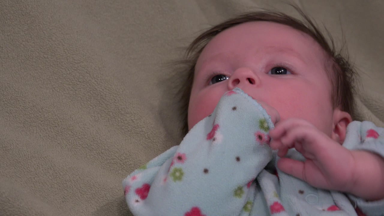 The baby remains awake on the tan bed in Westminster, Colorado, with one hand curled beside a pale blue patterned cloth covering part of the mouth.