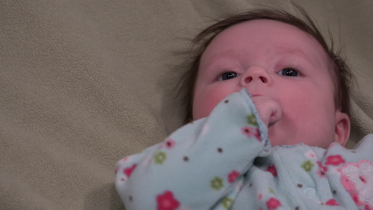 In Westminster, Colorado, a baby lies on a beige soft surface and looks upward while a pale blue floral sleeve rests near the mouth at the start of the clip.