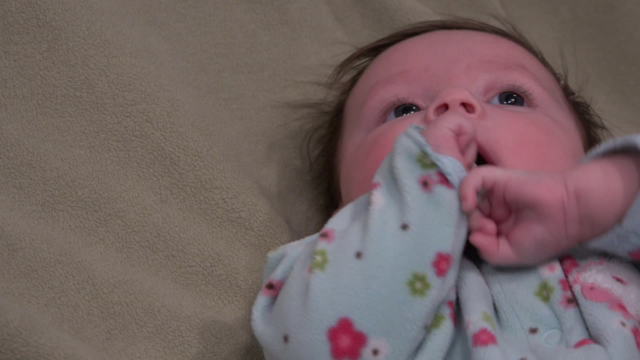 Baby lying on a beige bed with one hand near the mouth.