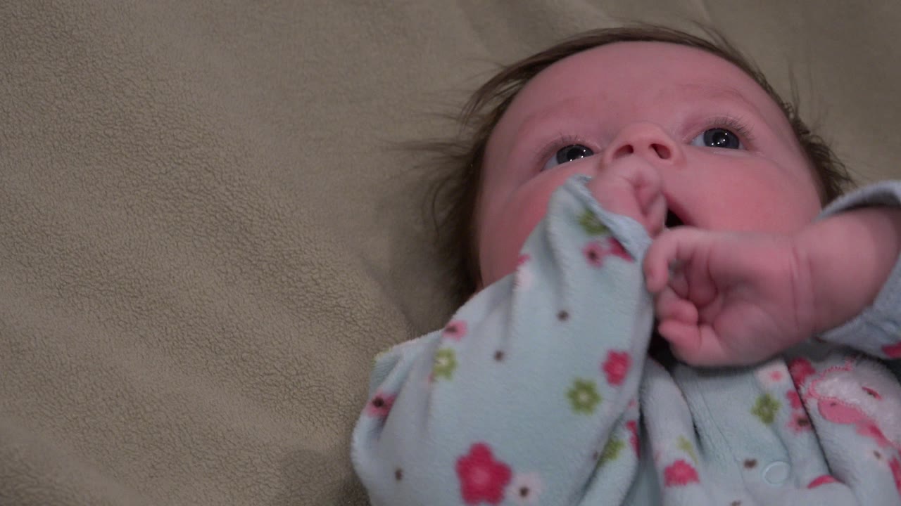 At 0:01 in Westminster, Colorado, the baby looks upward while both hands lift toward the face against the beige bed background.