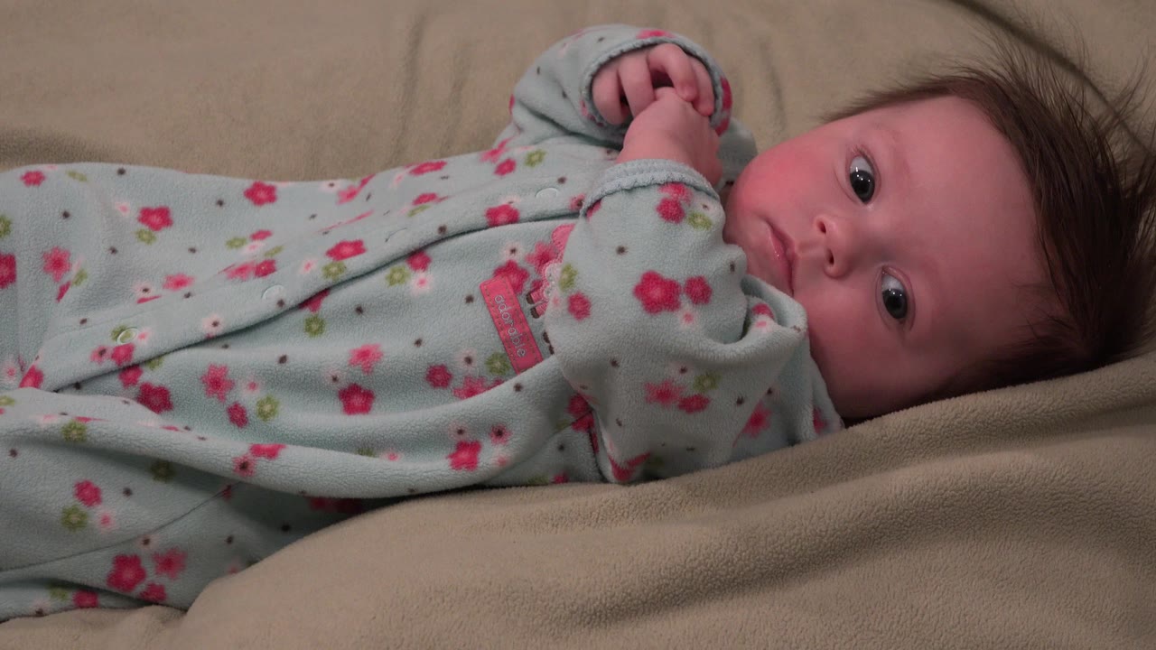 At 0:00, a young infant in Westminster, Colorado lies on beige bedding in a pale blue floral sleeper, looking toward the camera with hands near the chest.