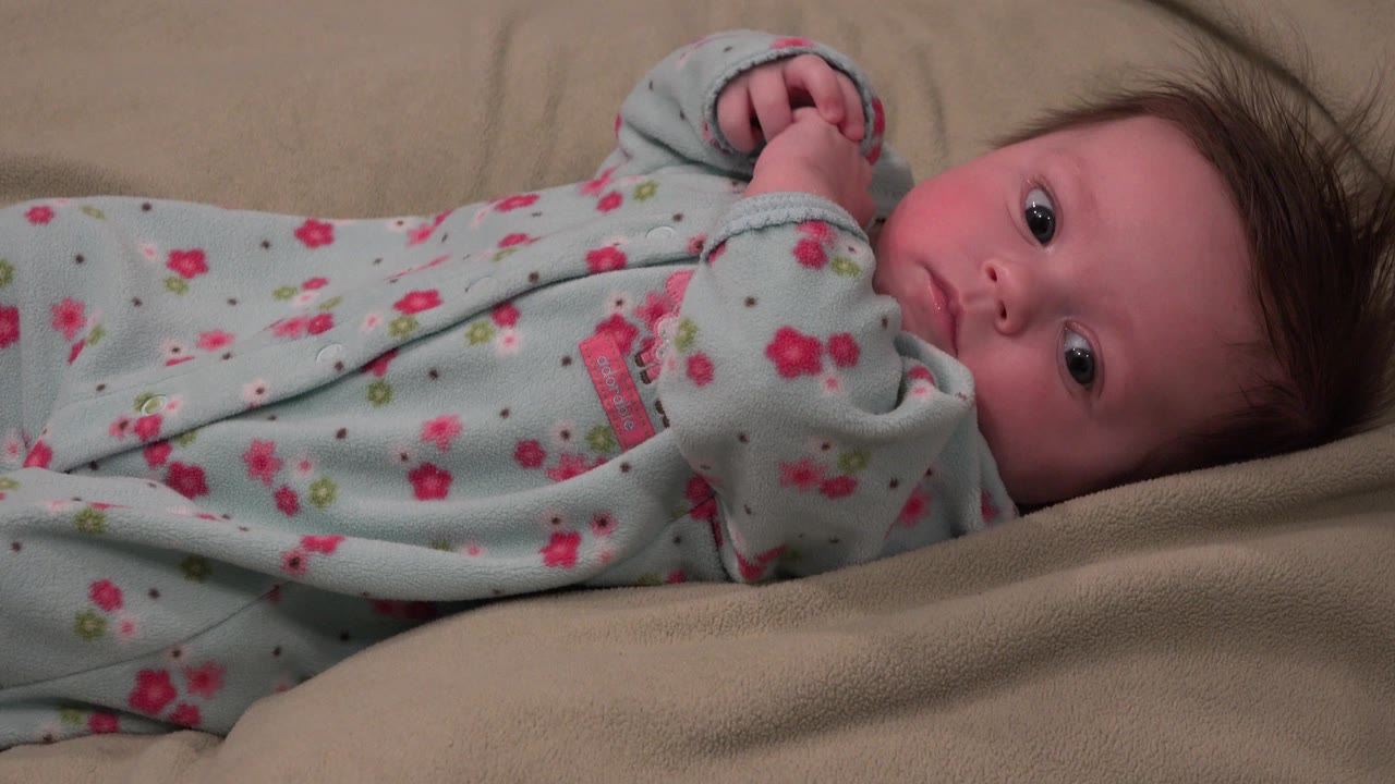At 0:01, the infant remains on their back in Westminster, Colorado, with both hands lifted near the face and the floral sleeper spread across the beige surface.