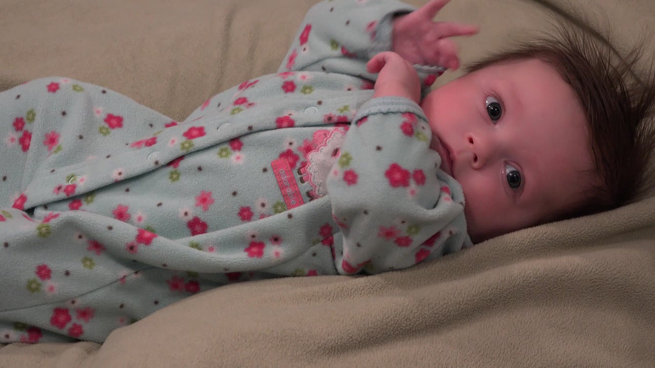 Baby in a floral sleeper lying on a beige blanket and looking toward the camera.