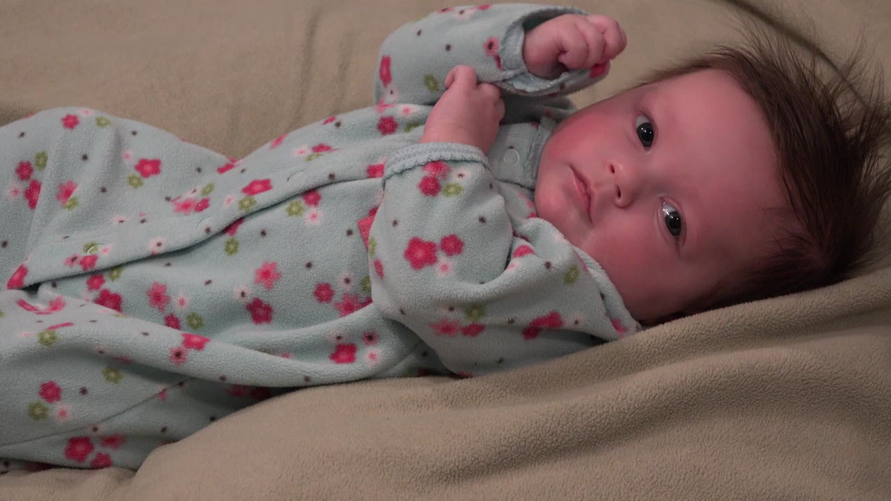 At 0:00, an awake baby in Westminster, Colorado lies on beige bedding in a pale blue floral sleeper, with one hand near the face and eyes open toward the camera.