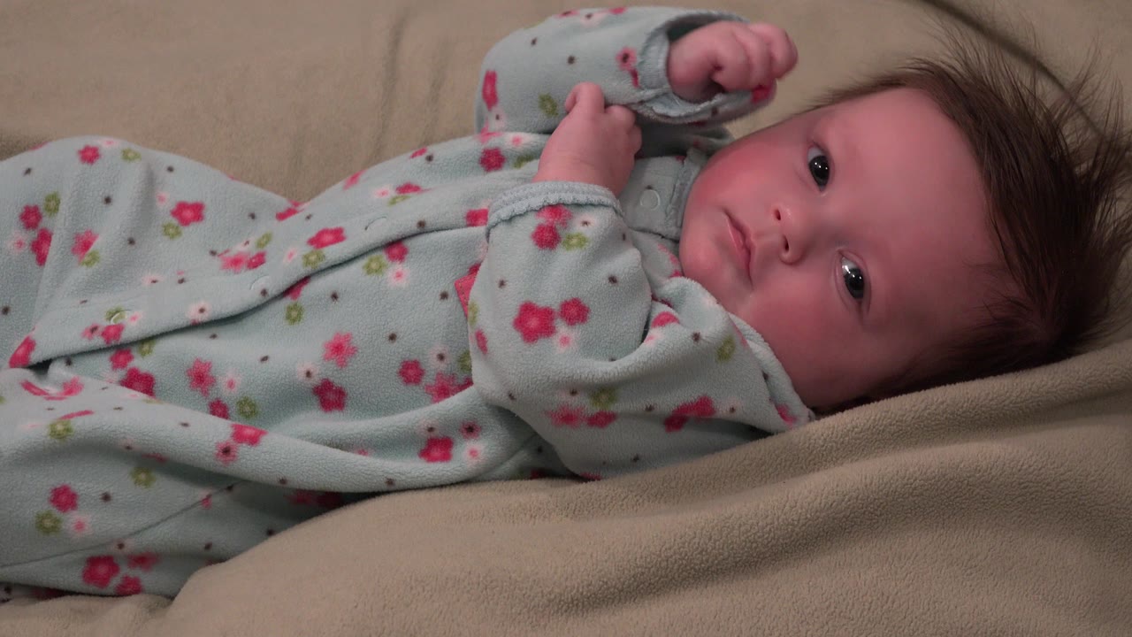 At 0:01, the baby remains on the beige cushion in Westminster, Colorado, looking toward the camera while the floral sleeves frame the cheeks and upper body.
