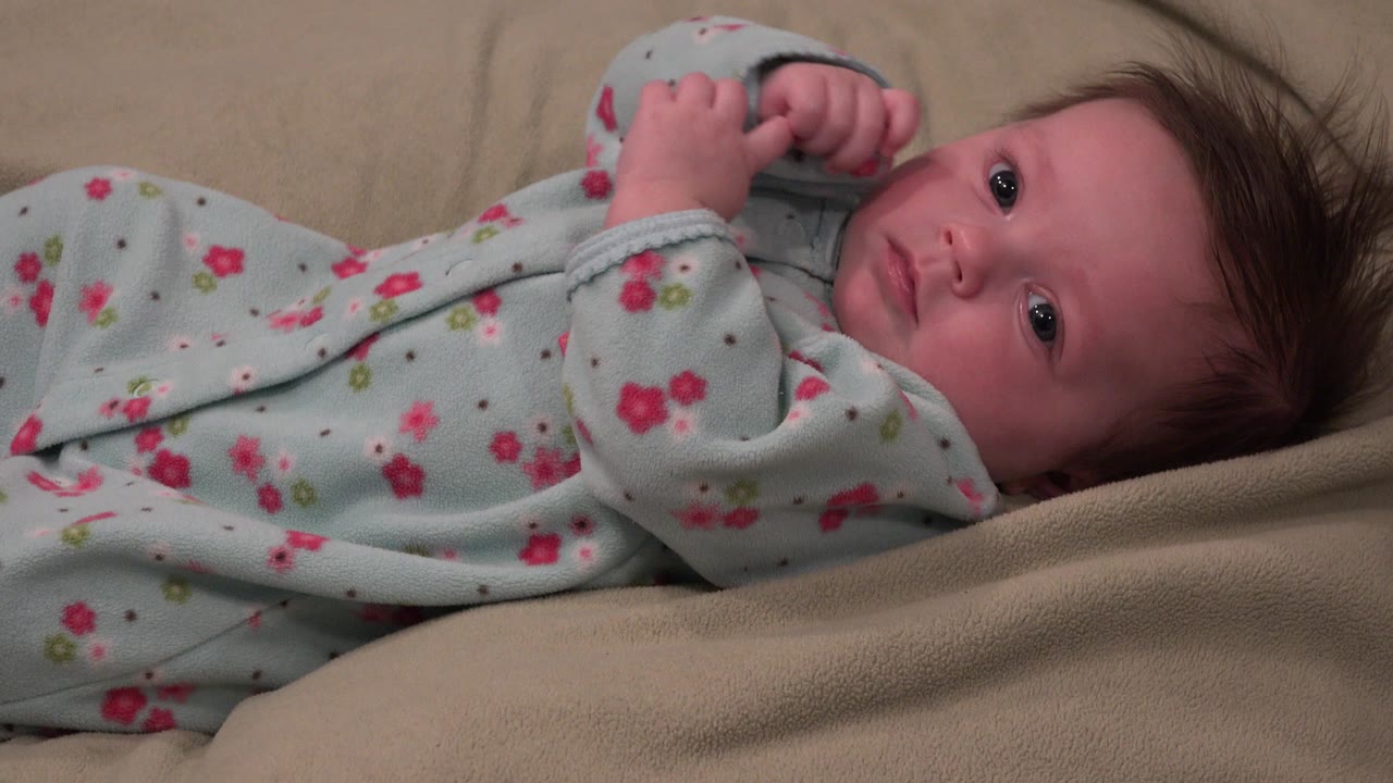 Baby in blue floral clothes lying on a beige blanket.