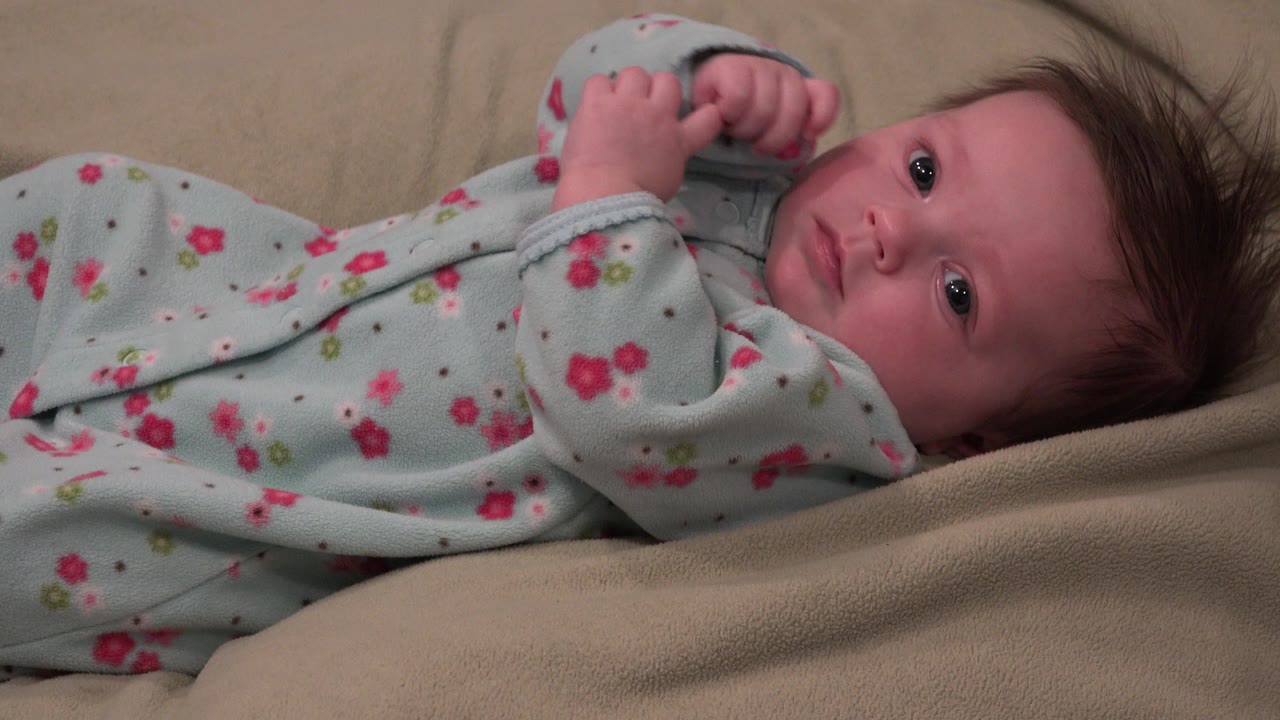 At 0:01 in Westminster, Colorado, the baby looks toward the camera while lying sideways across the beige blanket, with one sleeve bunched near the hands.
