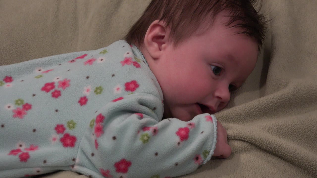 Baby in a pale blue floral sleeper lies belly down on a tan blanket in Westminster, Colorado, with the head turned toward the right edge of the frame.
