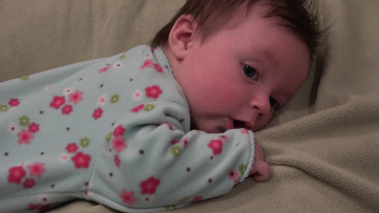 At 0:01, the baby remains on their stomach on the tan couch in Westminster, Colorado, with one eye visible and the sleeve pressed near the mouth.