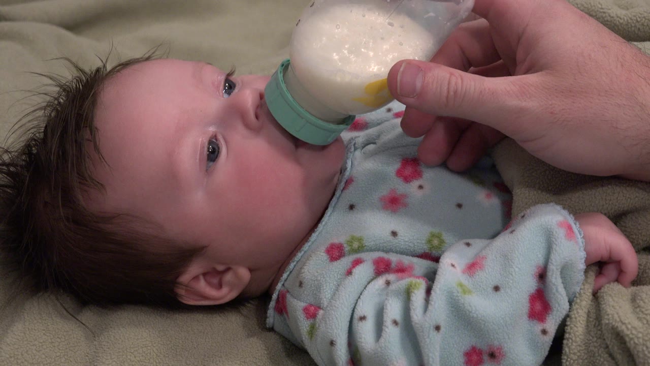 At 0:00 in Westminster, Colorado, a baby lies on beige bedding while an adult hand holds a milk bottle to the baby's mouth.