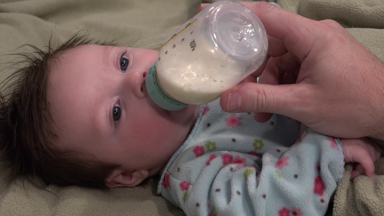 In Westminster, Colorado, a blue-eyed baby lies on a beige blanket while an adult hand holds a milk bottle at the baby's mouth at the start of the clip.