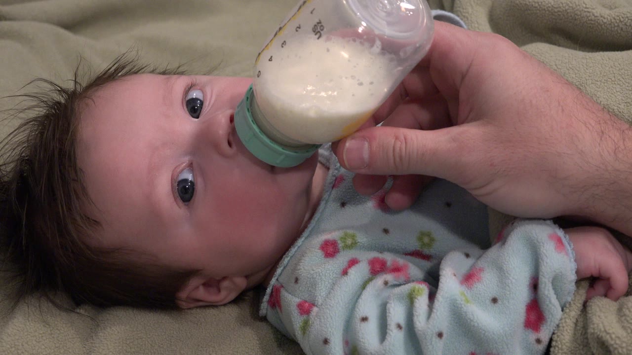 Infant lying on a blanket while drinking from a bottle held by an adult hand.